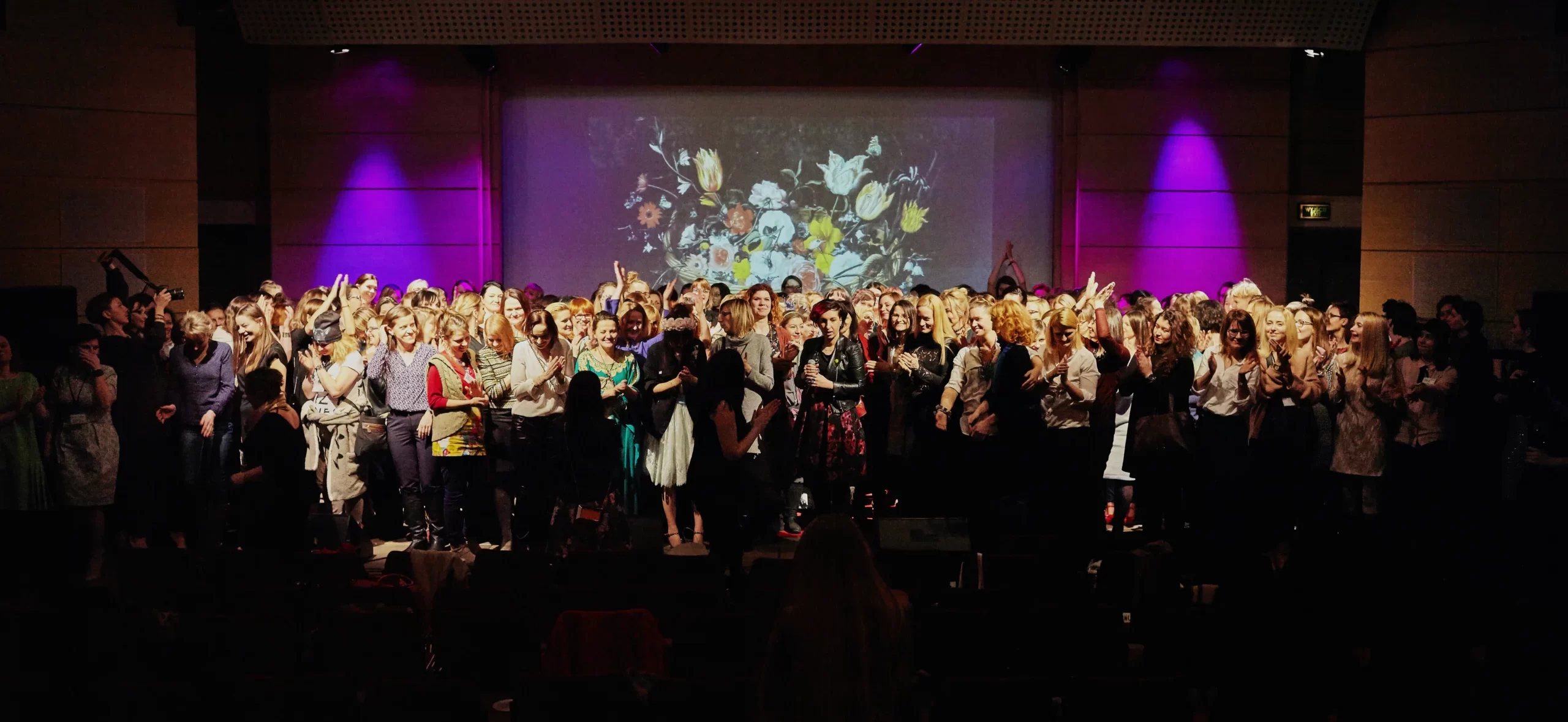 Zdjęcie na którym jest bardzo dużo kobiet stojących na scenie w tele jest ekran na którym wyświetlone są kwiaty.
