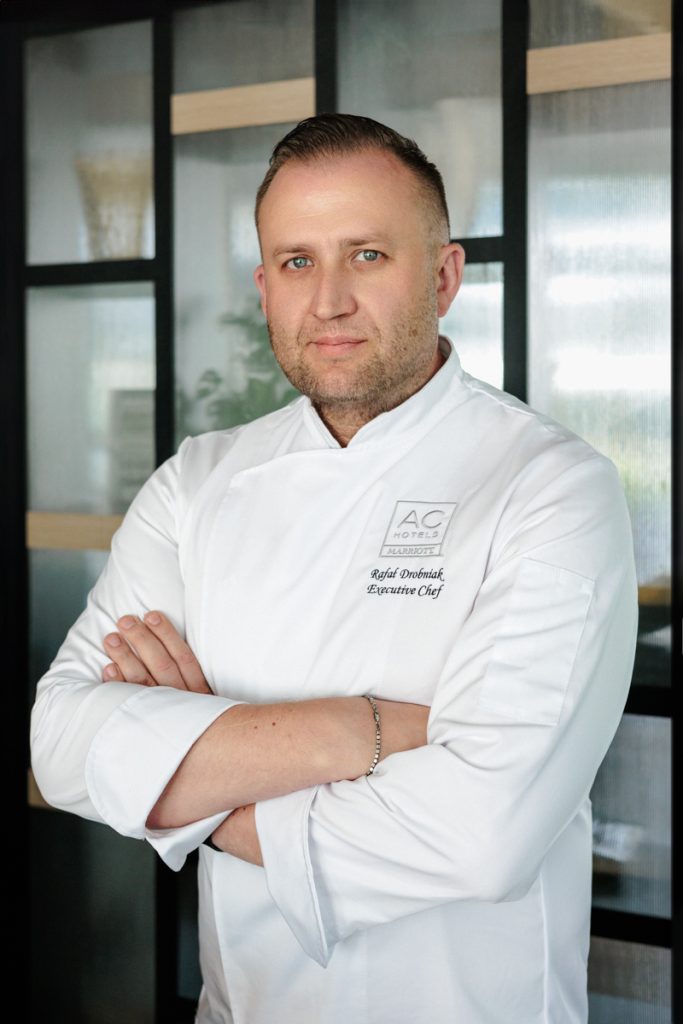 mężczyzna z założonymi rękami, w fartuchu kucharskim, na przeszklonym tle